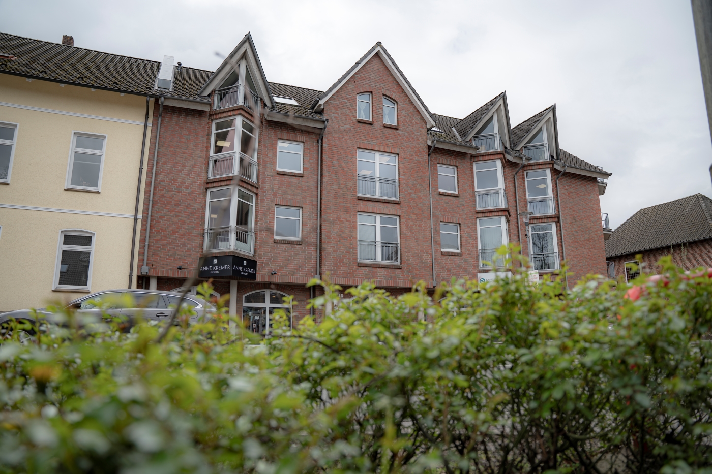 Eine braune Hausfassade. Im Vordergrund sind Büsche zu sehen.