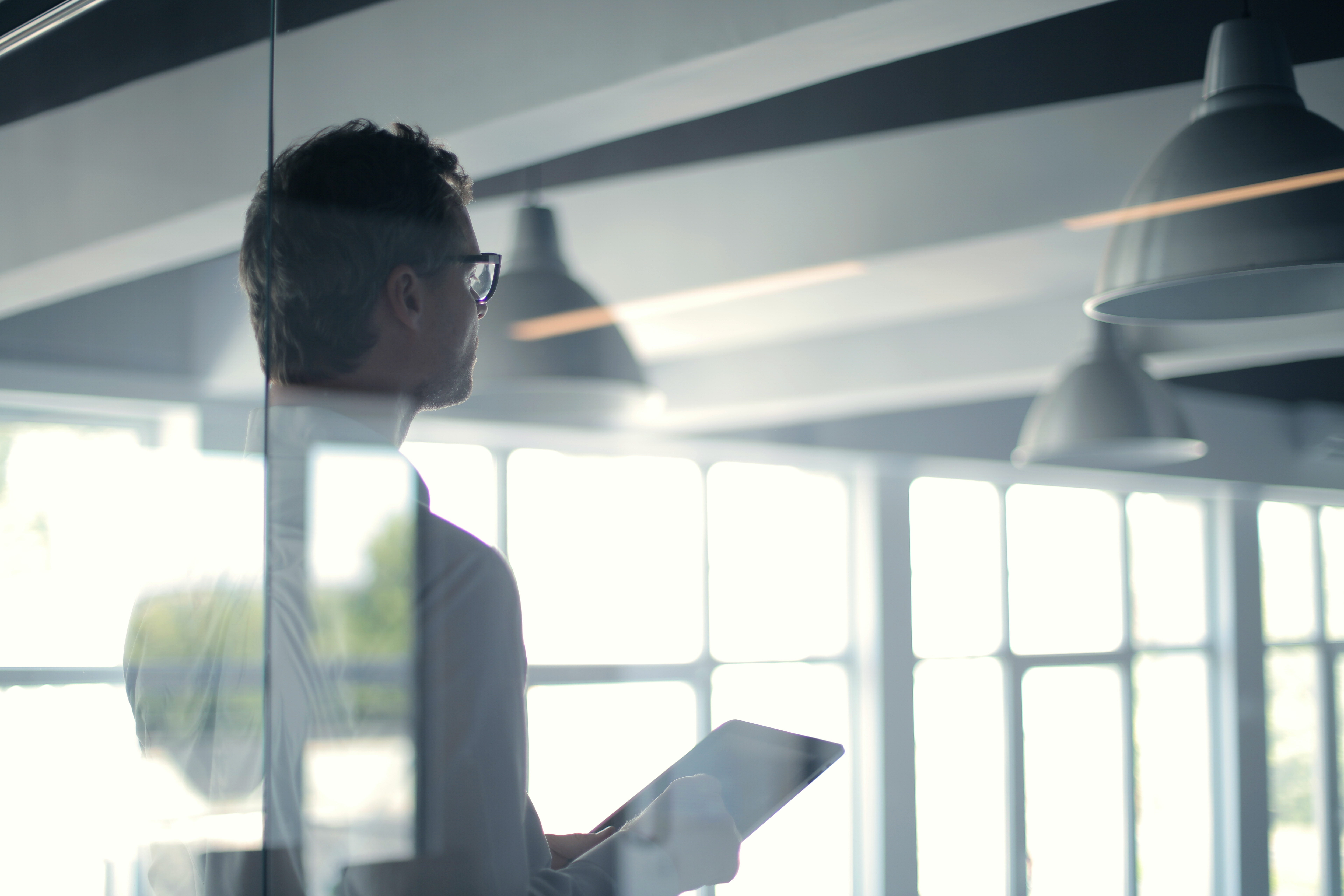 Ein Mann mit Brille steht seitlich gedreht hinter einer Glaswand in einem industriellen Büro und hält ein Tablet in der Hand.