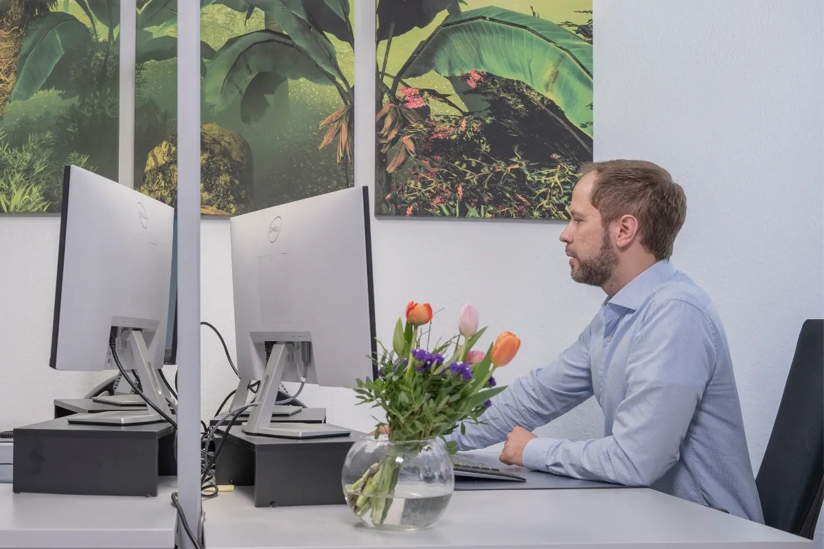 Ein Mann mit hellblauem Hemd sitzt an einem Schreibtisch und arbeitet, vor ihm steht ein Bildschirm.