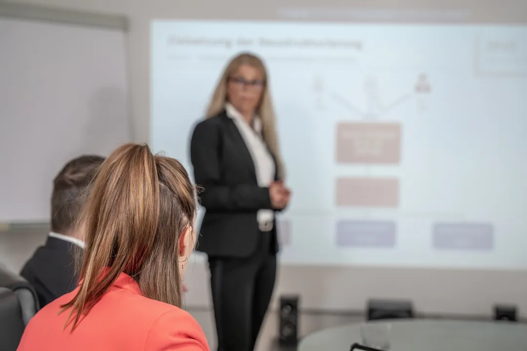 Mehrere Personen sind in einem Meetingraum. Eine Frau mit Brille steht im Hintergrund vor einer Leinwand, auf der eine Präsentation zu sehen ist.