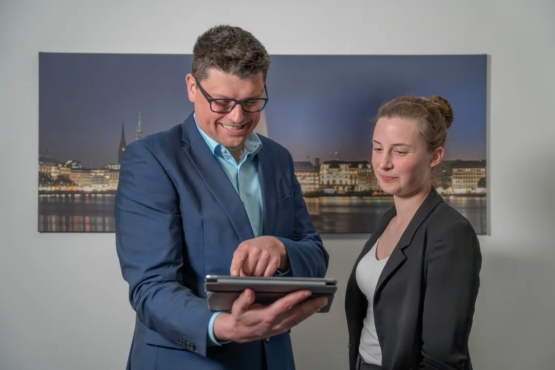 Ein Mann und eine Frau stehen nebeneinander. Der Mann hält ein Tablet in der Hand und zeigt der Frau darauf etwas.