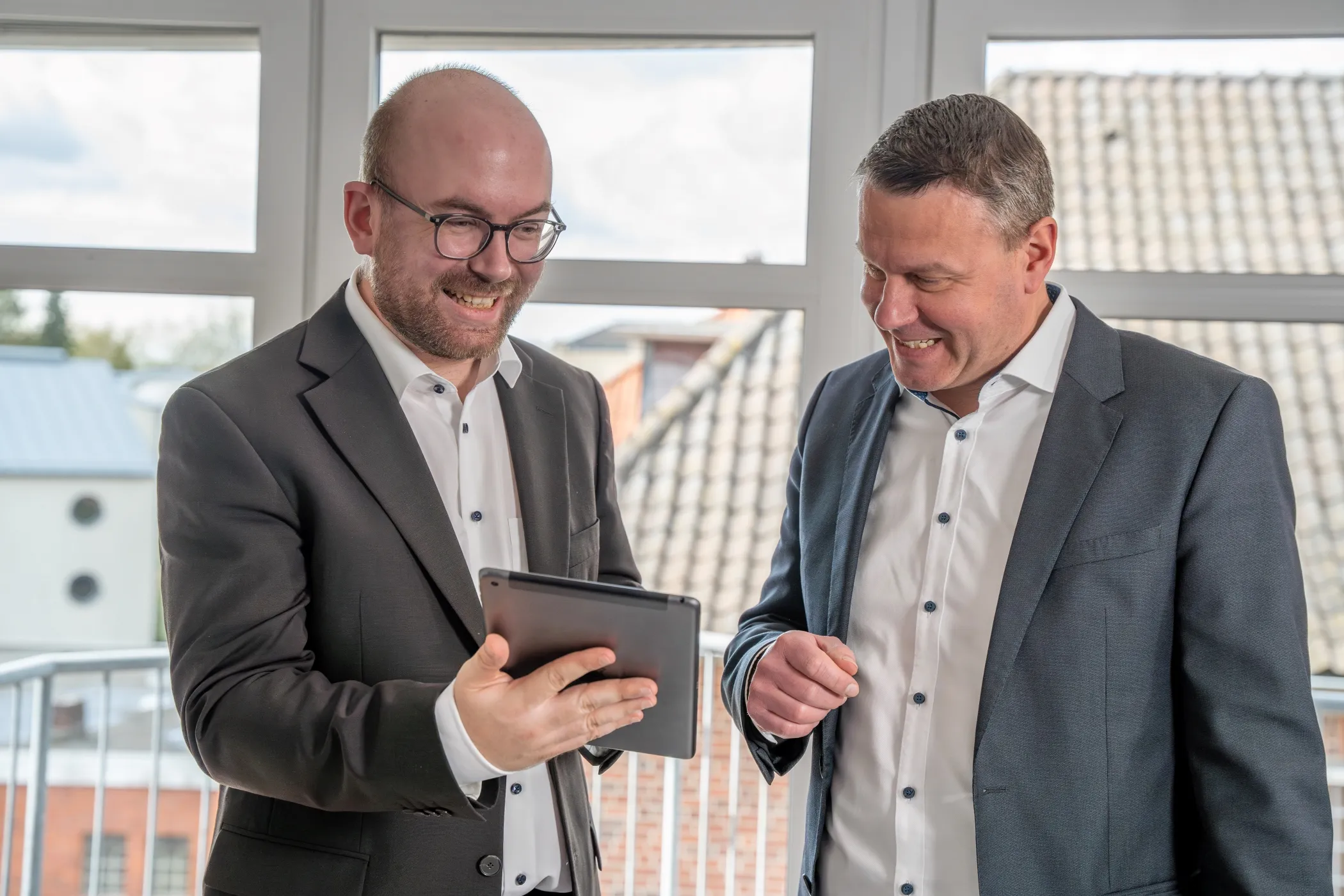 Zwei Männer stehen nebeneinander vor einem Fenster. Der Mann hält ein Tablet in der Hand und zeigt darauf etwas.