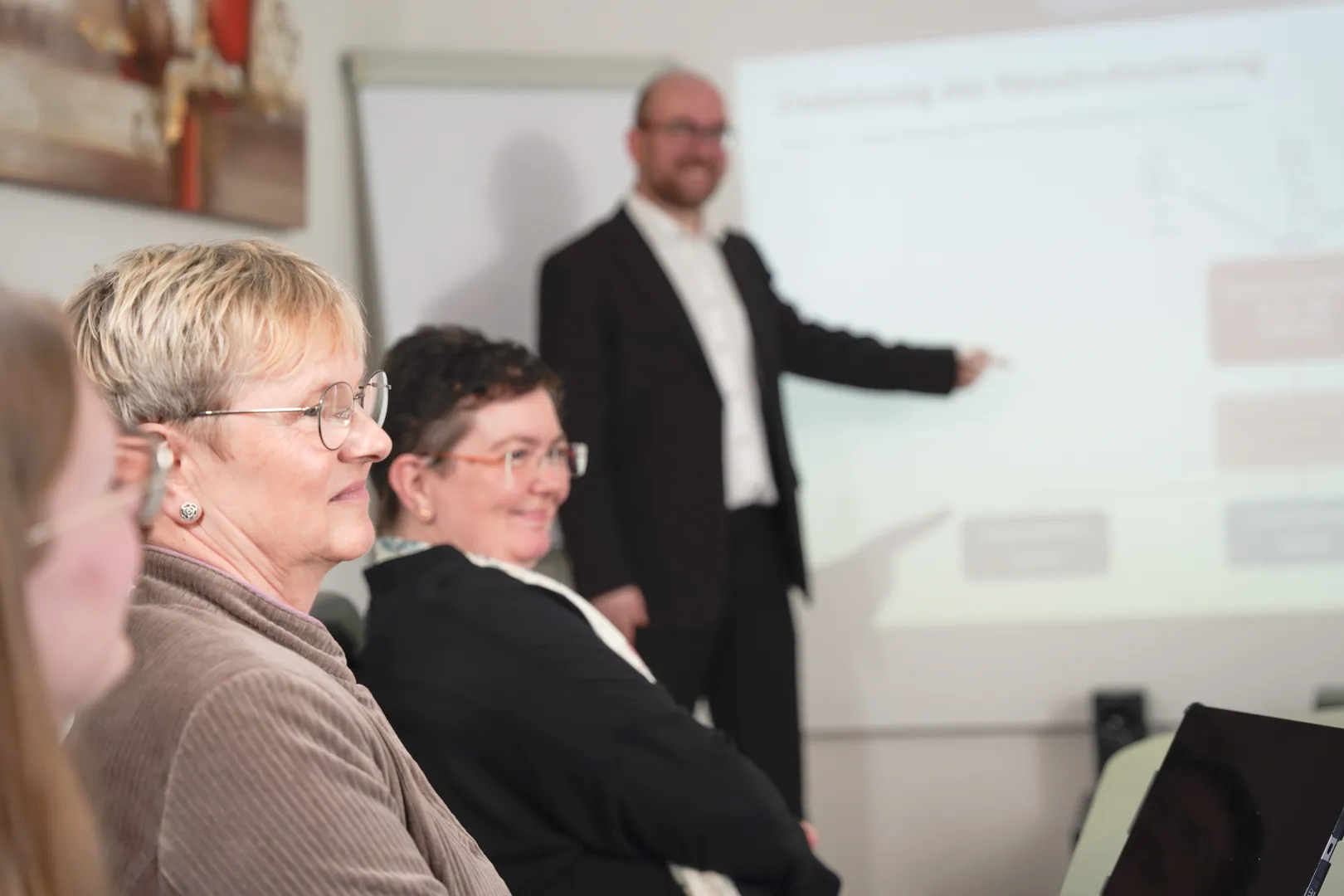 Mehrere Personen sitzen an einem Konferenztisch und ein Mann steht vor einer Leinwand, auf der eine Präsentation gezeigt wird.