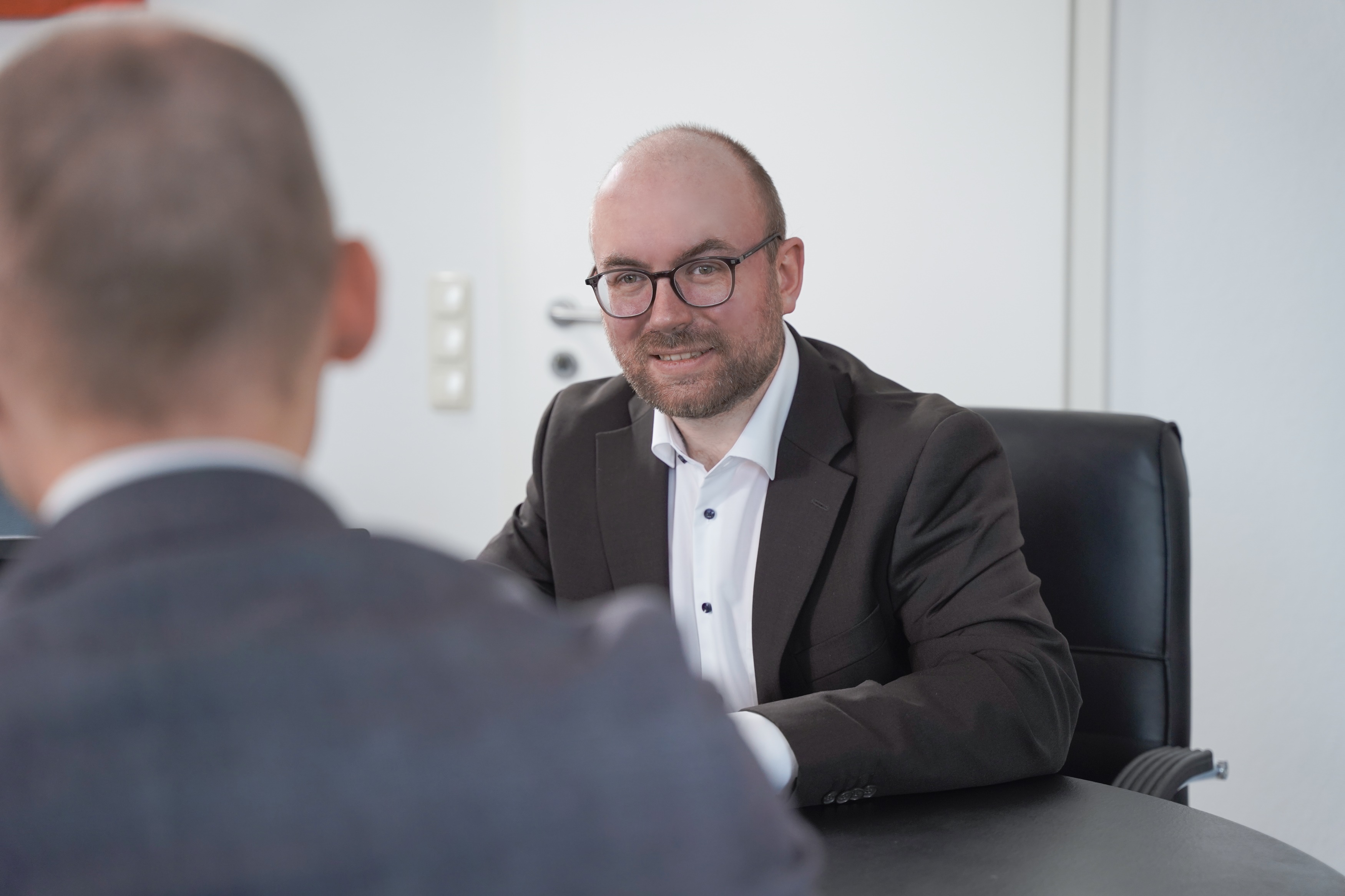 Eiin Mann mit Glatze und Brille sitzt an einem Schreibtisch. Von hinten sieht man einen anderen Mann. Sie sprechen miteinander.
