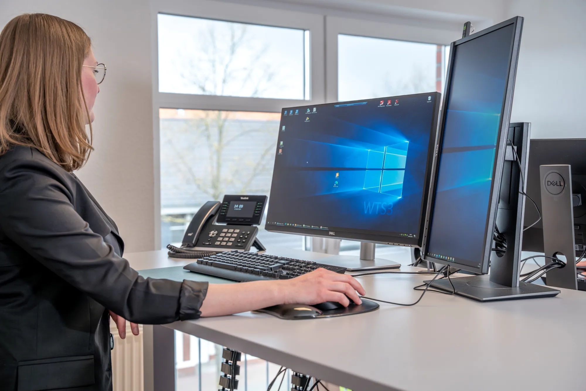 Eine Frau steht seitlich an einem Schreibtisch und arbeitet an zwei Bildschirmen.