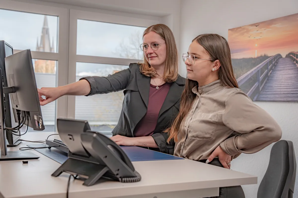 Zwei junge Frauen stehen an einem Schreibtisch. Die Frau auf der linken Seite zeigt mit einer Hand auf einen Bildschirm, der vor ihr steht.