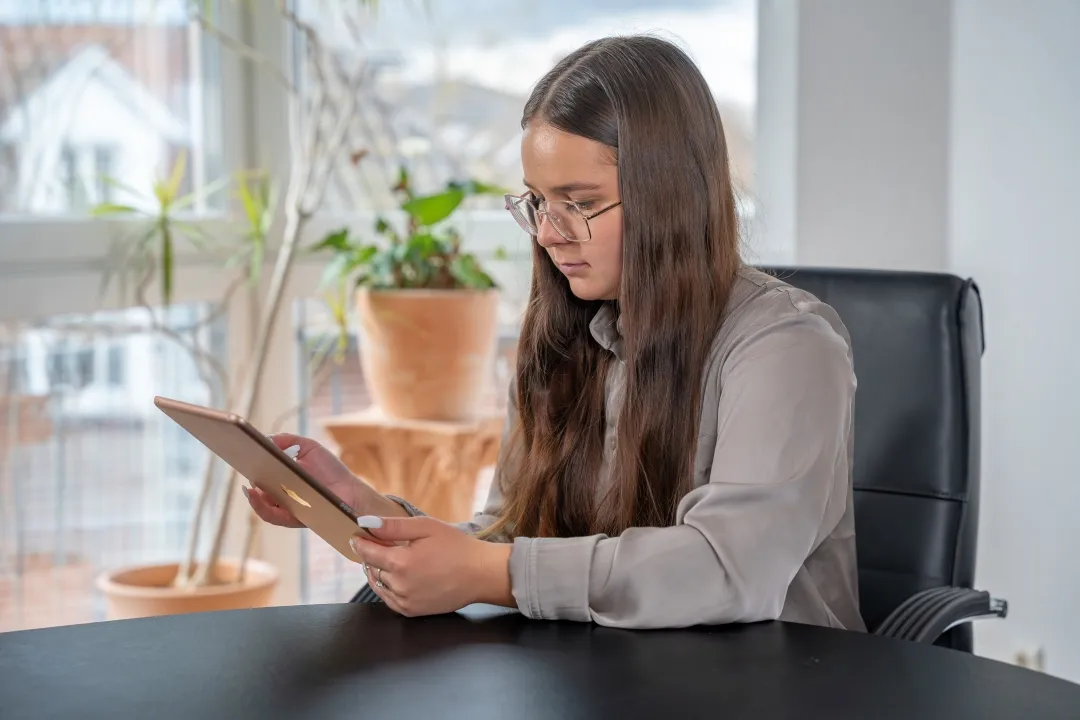 Eine junge Frau mit Brille sitzt an einem Schreibtisch und schaut auf ein Tablet, das sie in der Hand hält.