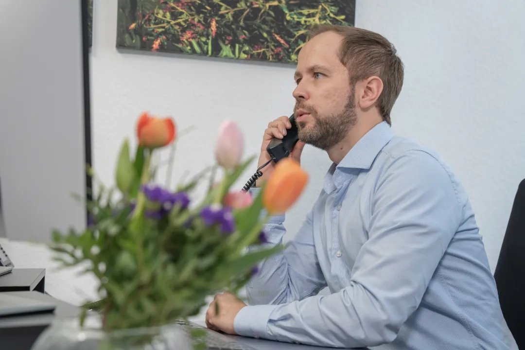 Ein Mann mit einem blauen Hemd sitzt an einem Schreibtisch und telefoniert.