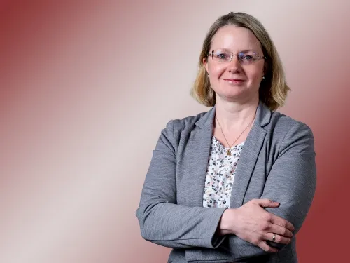 Porträt einer blonden Frau mit Brille und grauem Blazer.