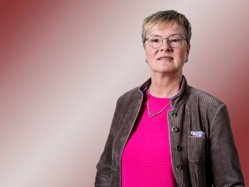 Porträt einer blonden Frau mit Brille und pinken T-Shirt sowie braunen Blazer.