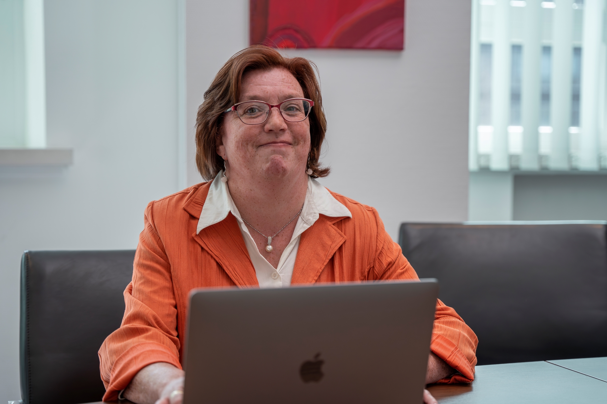 Eine Frau mit einem orangen Blazer sitzt mit einem Laptop an einem Tisch und lächelt in die Kamera.
