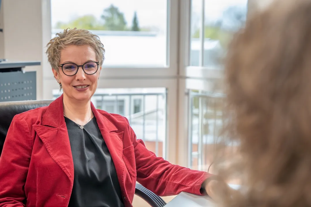 Eine Frau in einem roten Blazer sitzt auf einem Schreibtischstuhl und lächelt.